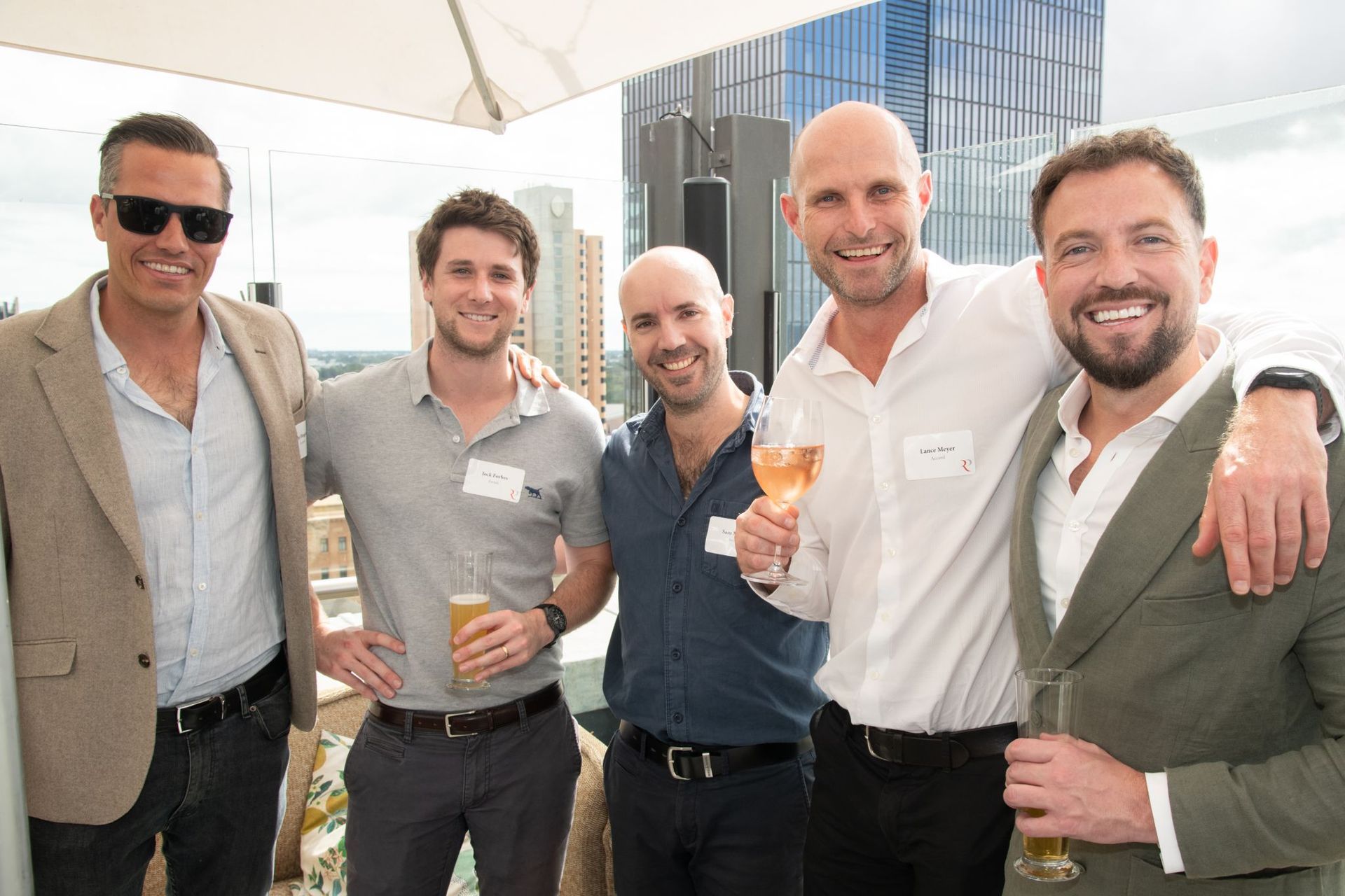 Five men in business casual attire smile while standing together with drinks at an outdoor rooftop event.