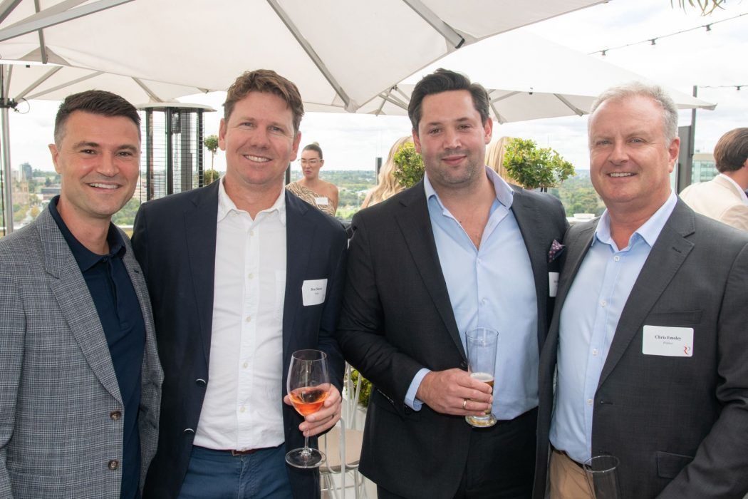 Four men in business casual attire smile while holding drinks at an outdoor rooftop event under white umbrellas.