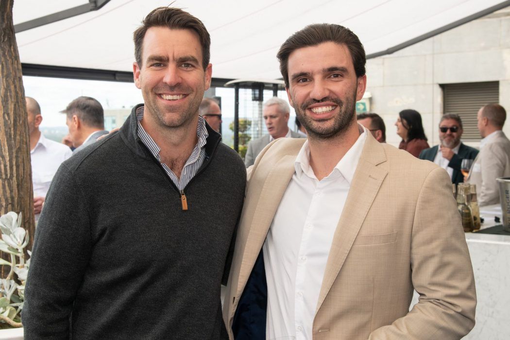 Two men stand smiling side-by-side at an outdoor gathering, one wearing a dark sweater and the other a beige blazer.