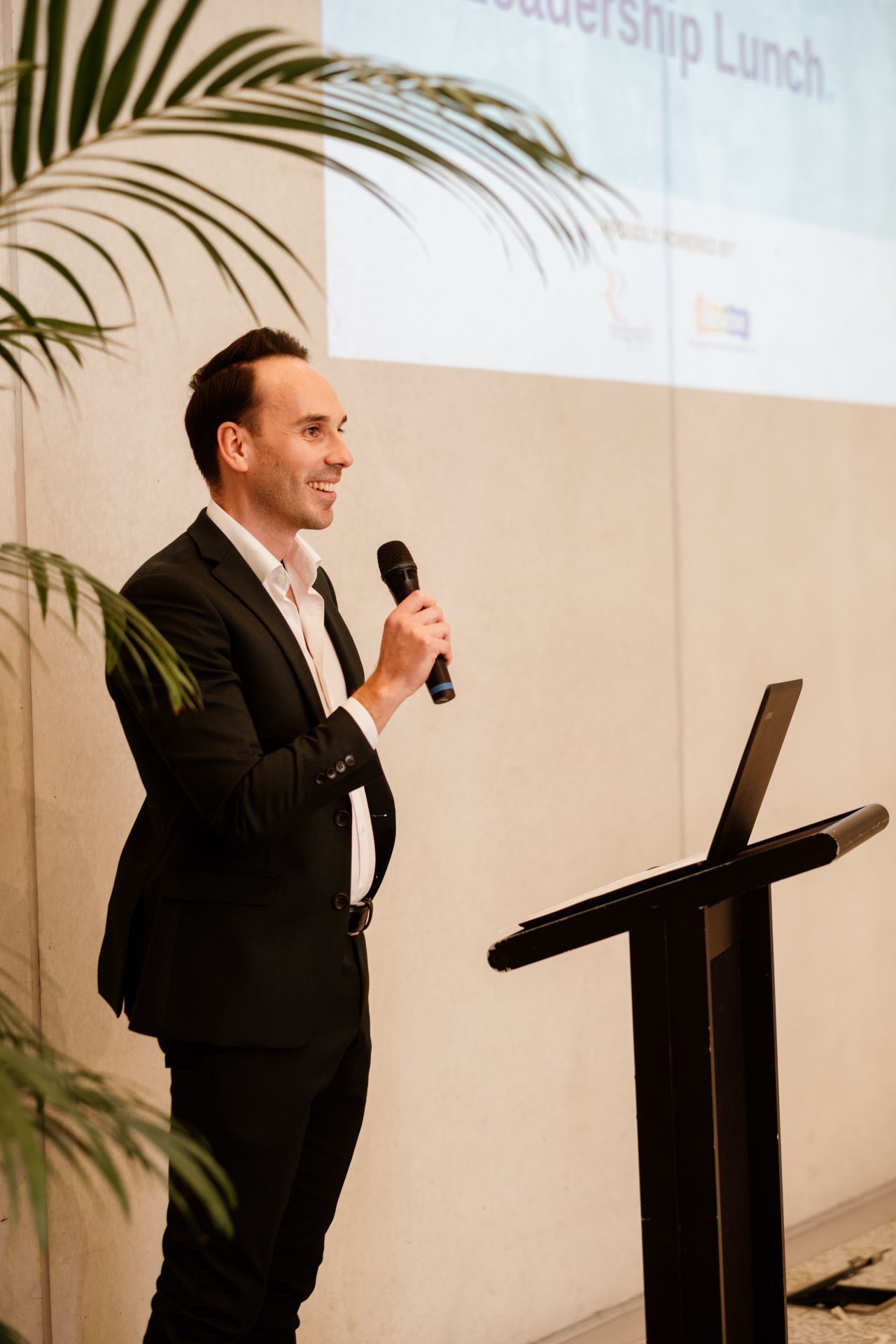 A professional in a suit speaks into a microphone at a podium during a leadership lunch event.