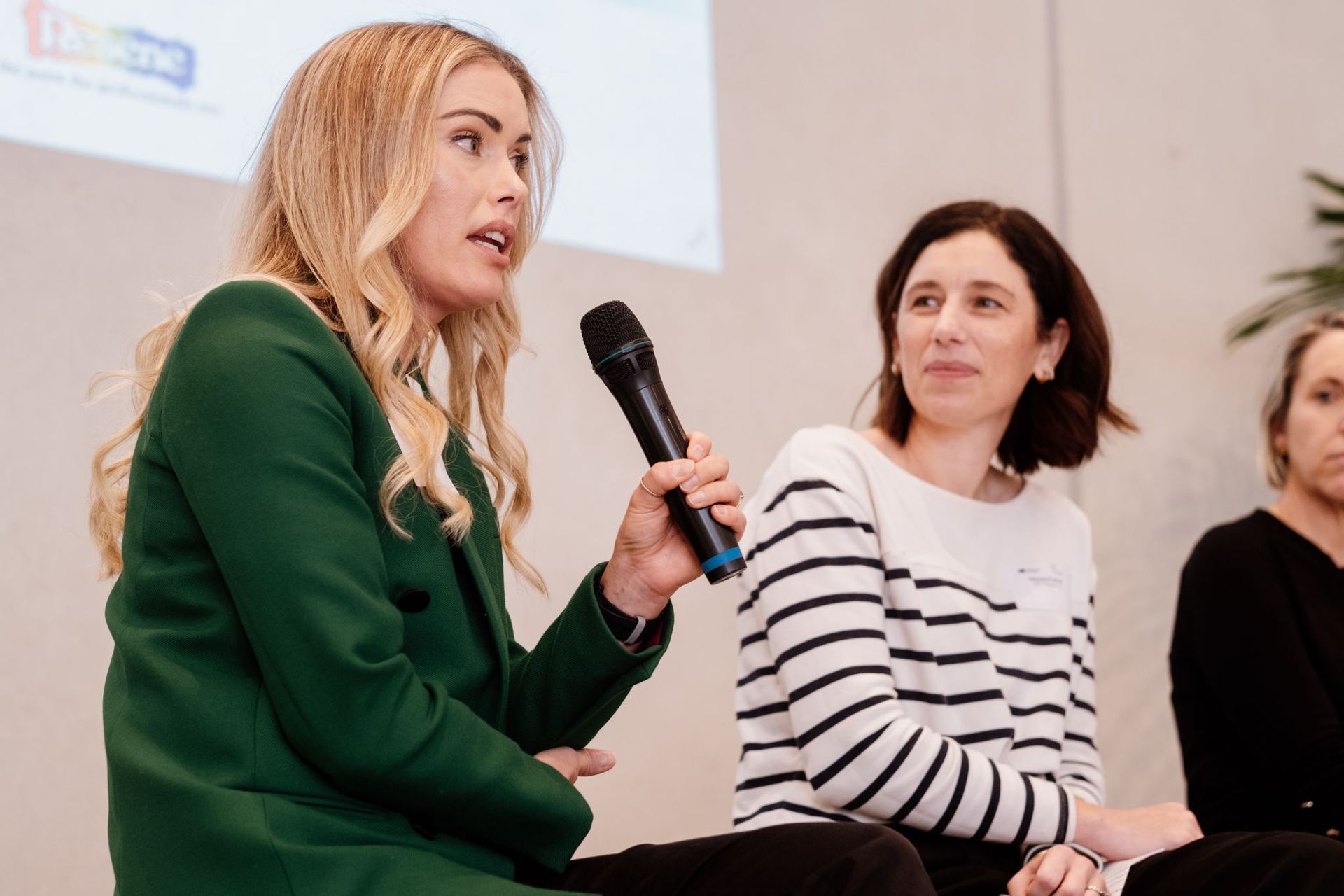 A person in a green blazer speaks into a microphone while two others listen during a presentation.