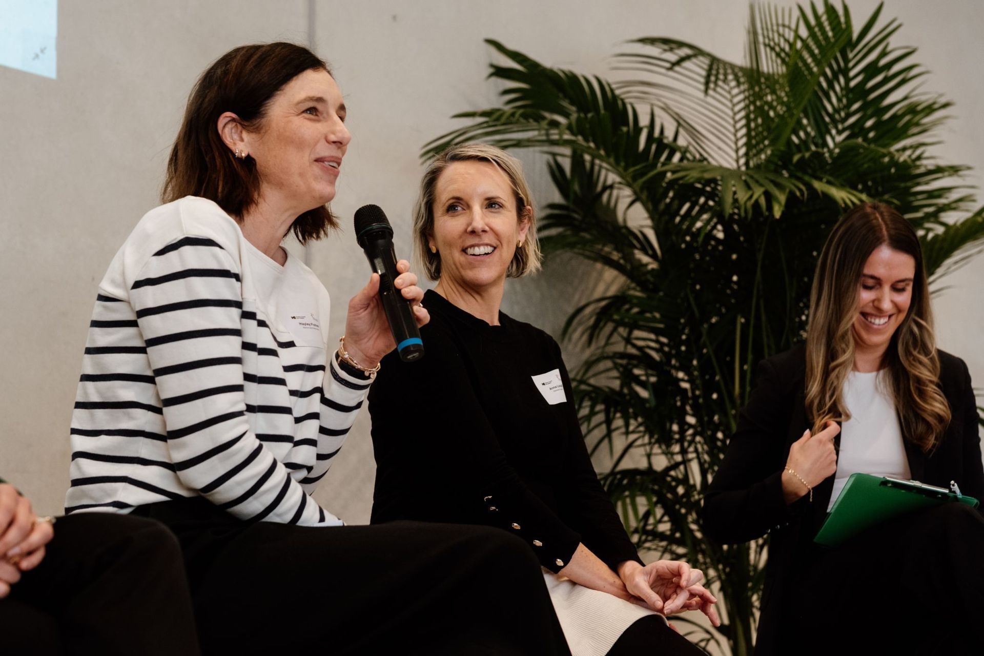 Three people sitting on a stage at an indoor event, one holding a microphone and speaking to an audience.