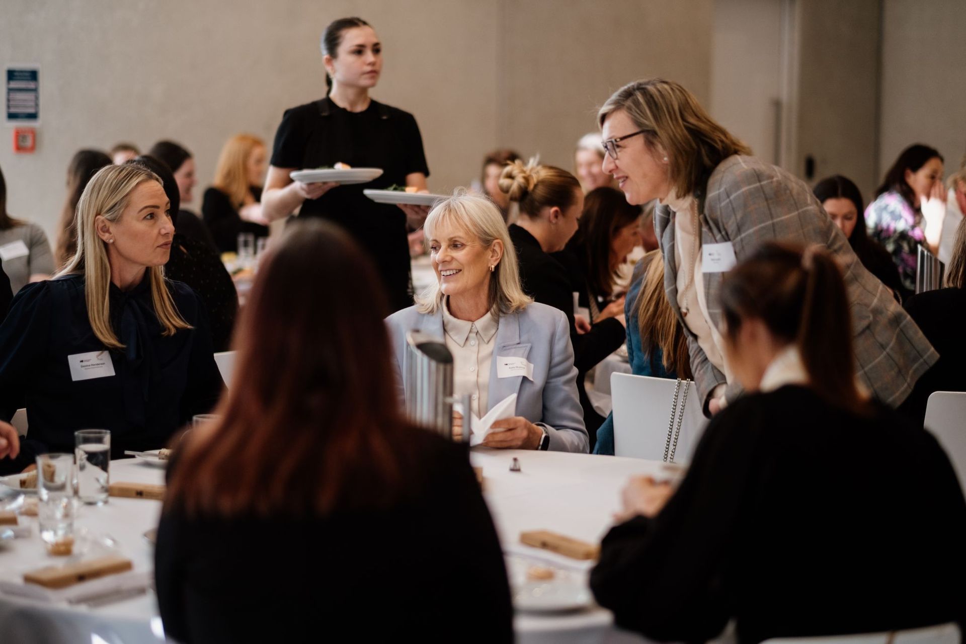 People sit at round tables in a conference room while a server holds plates and an attendee leans in to talk to a peer.
