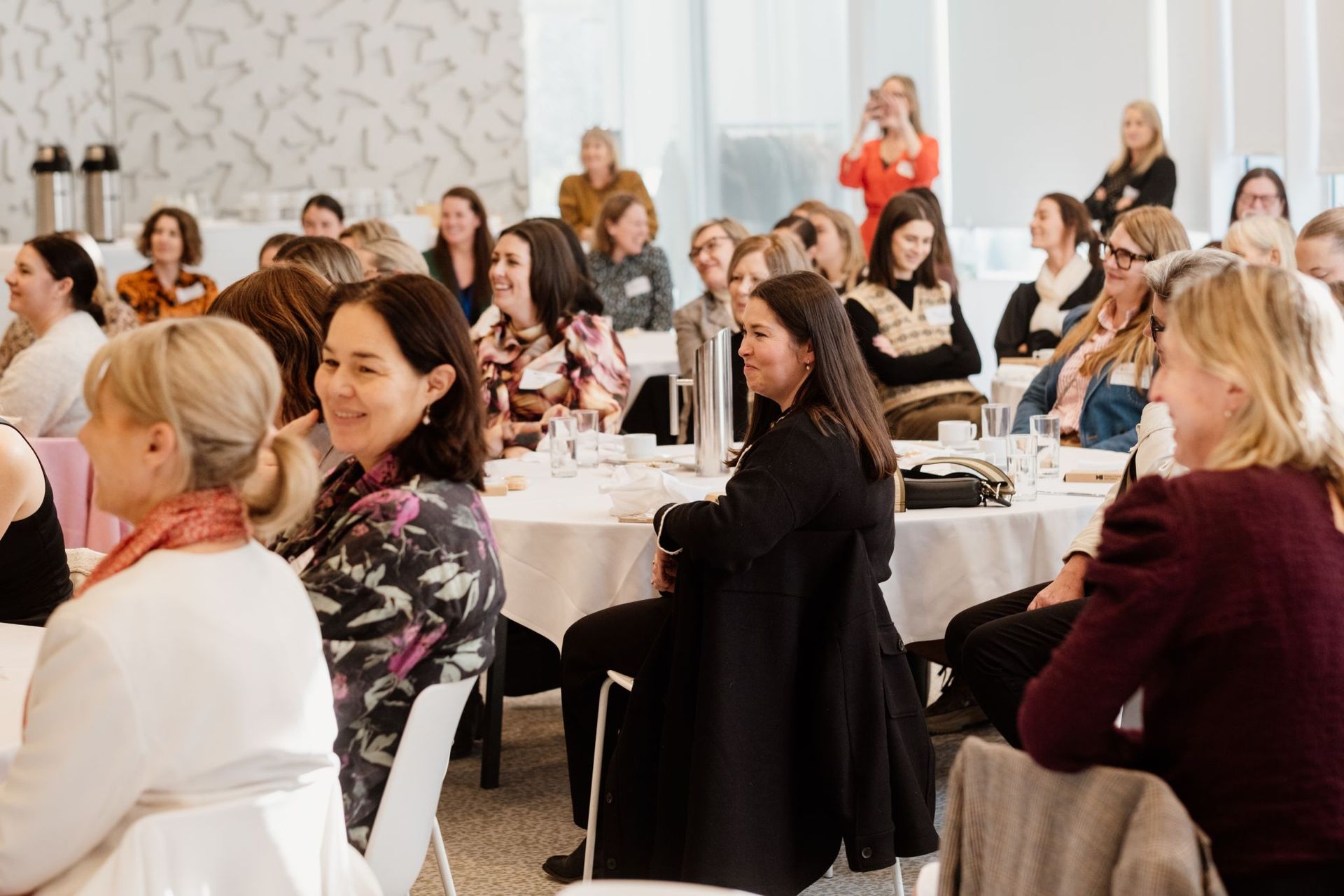 A group of people sitting at round tables in a bright, modern room during a professional conference or seminar.