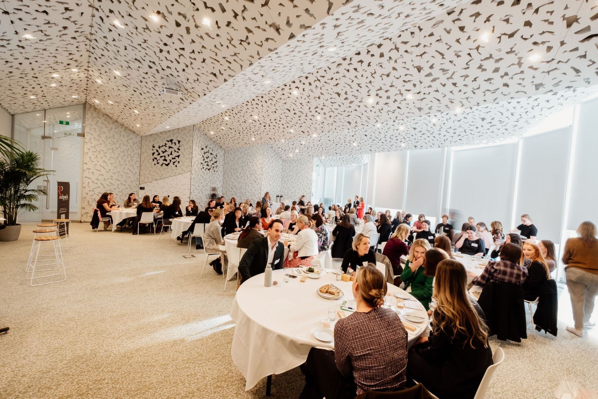 A large, bright event space with many people seated at round tables for a gathering under a unique, patterned ceiling.