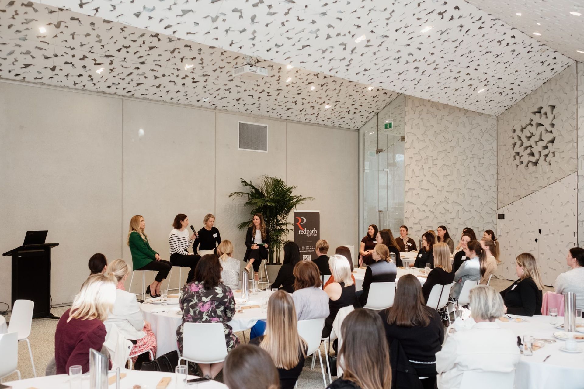 A panel of speakers addresses an audience in a bright, modern room with patterned ceilings and large indoor plants.