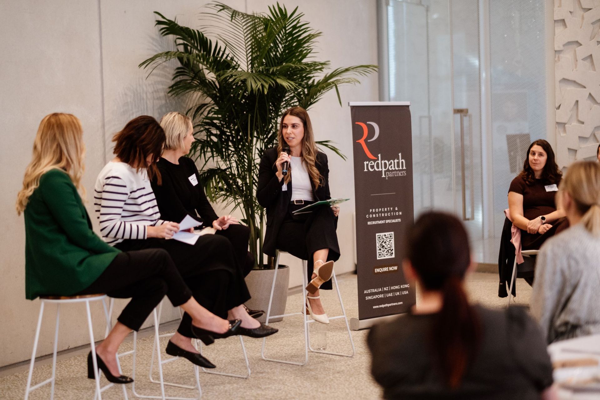 Four panelists sit on stools on stage, engaged in discussion near a banner for Redpath Partners, with an audience watching.