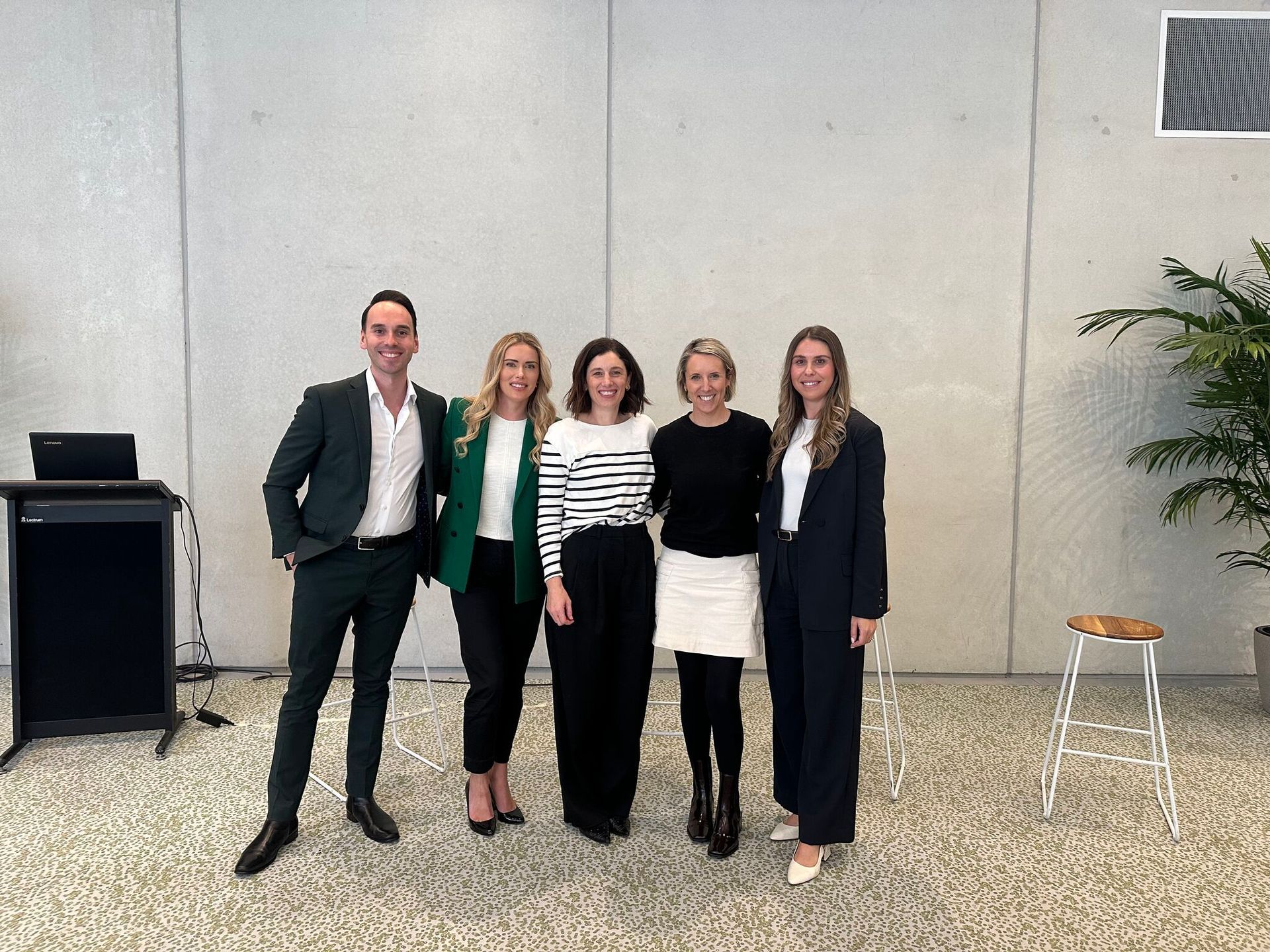 Five people stand in a line against a light grey wall in a bright, modern interior, posing for a group photo.