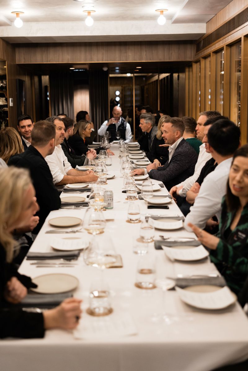 A large group of people sit around a long, white-clothed dining table in a warm, dimly lit restaurant.