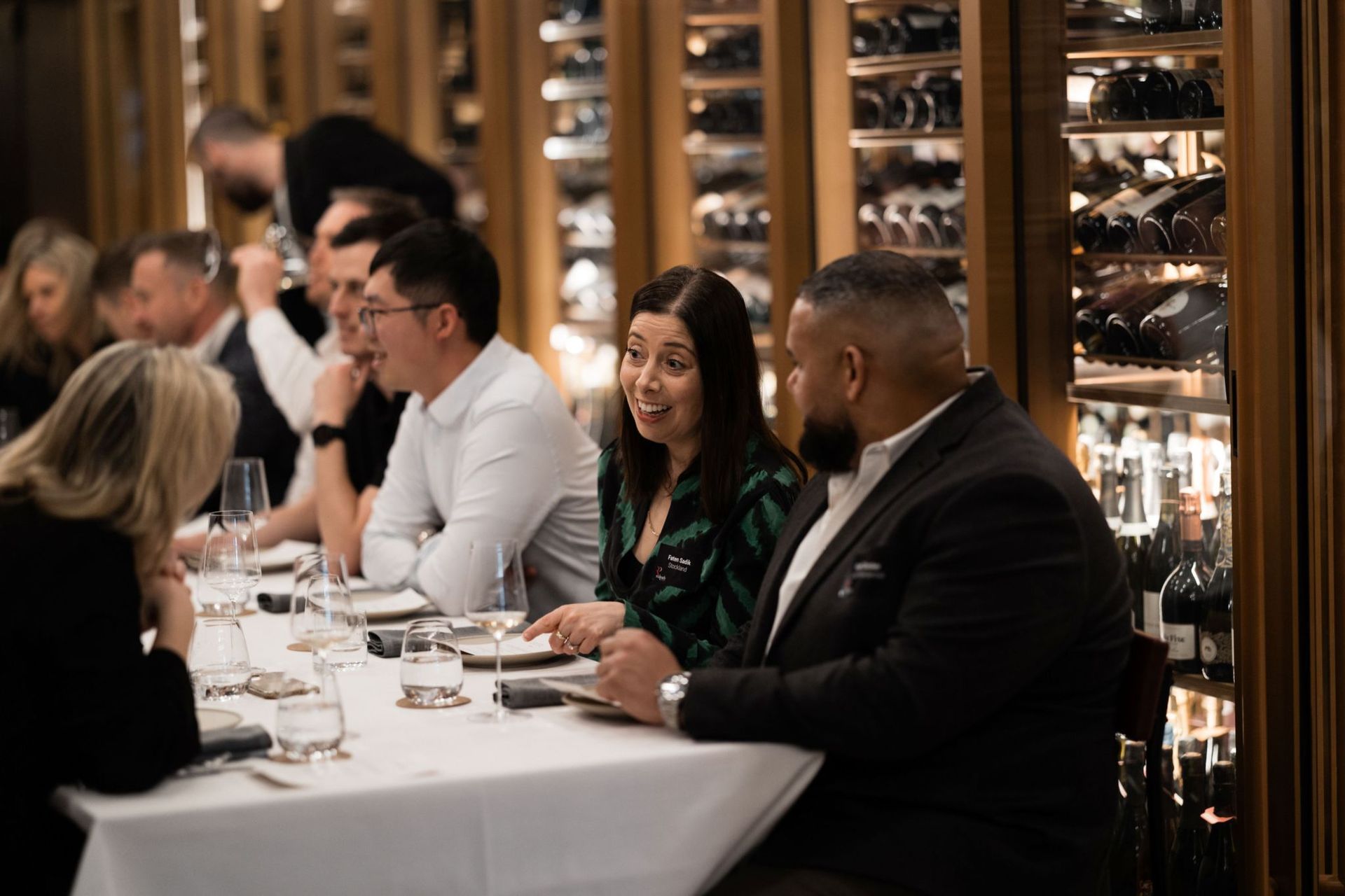 A group of people dining at a long table in a restaurant with wine racks in the background.
