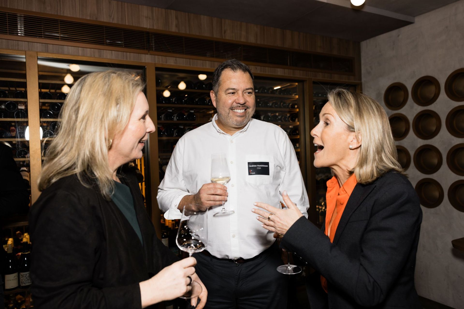 Three people in business attire hold glasses of wine and laugh together inside a well-lit restaurant or wine cellar.