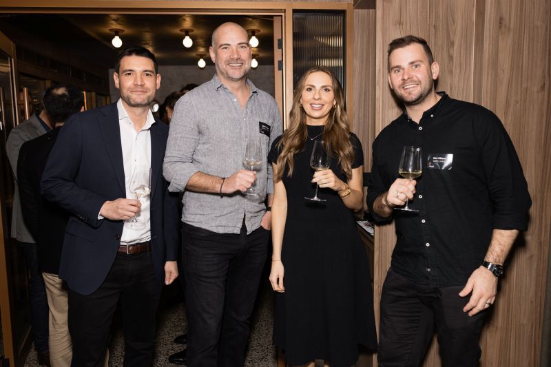Four people in business casual attire smile while holding wine glasses in a modern, warmly lit interior.