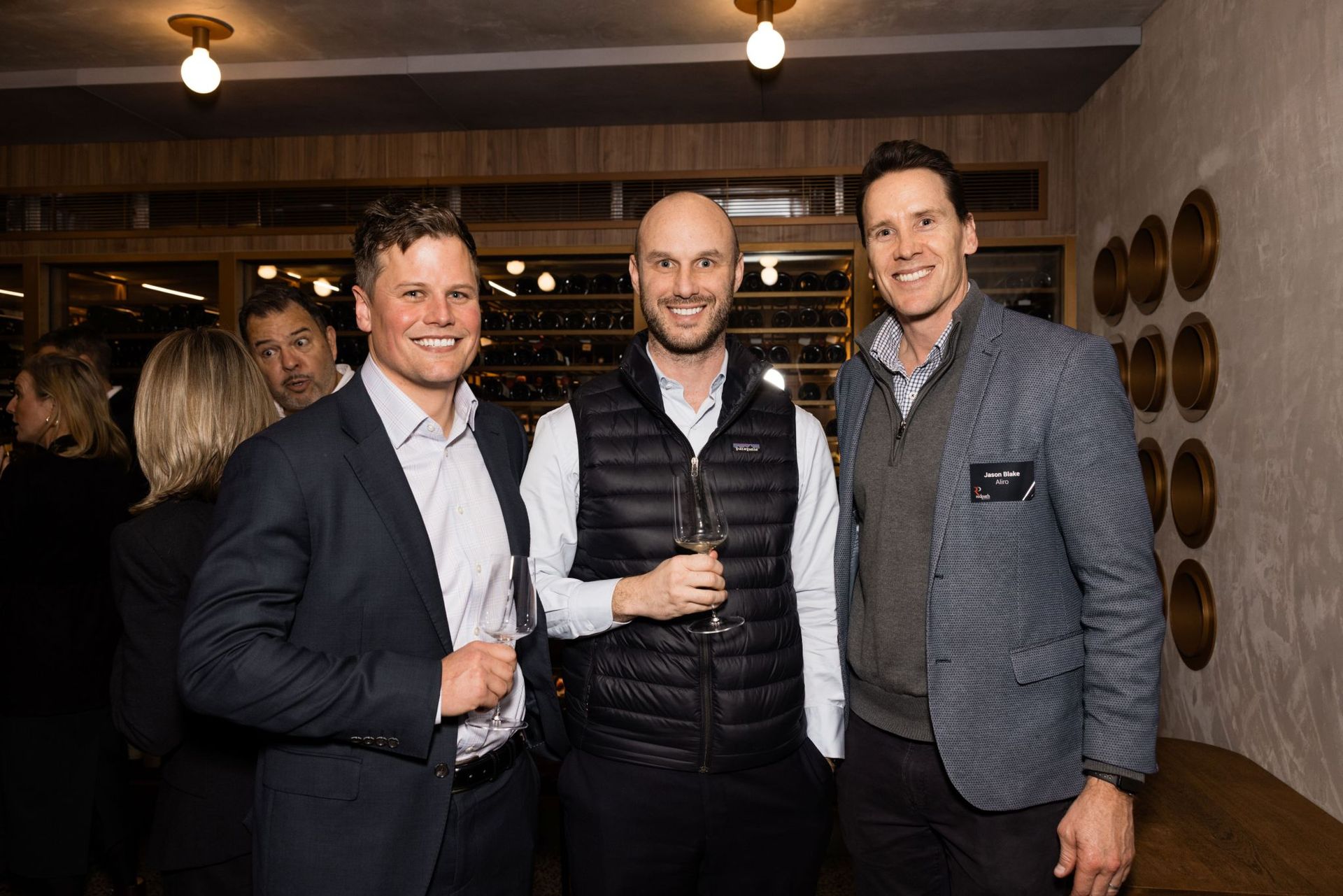Three men smiling, standing side-by-side in a restaurant or event space, with others blurred in the background.