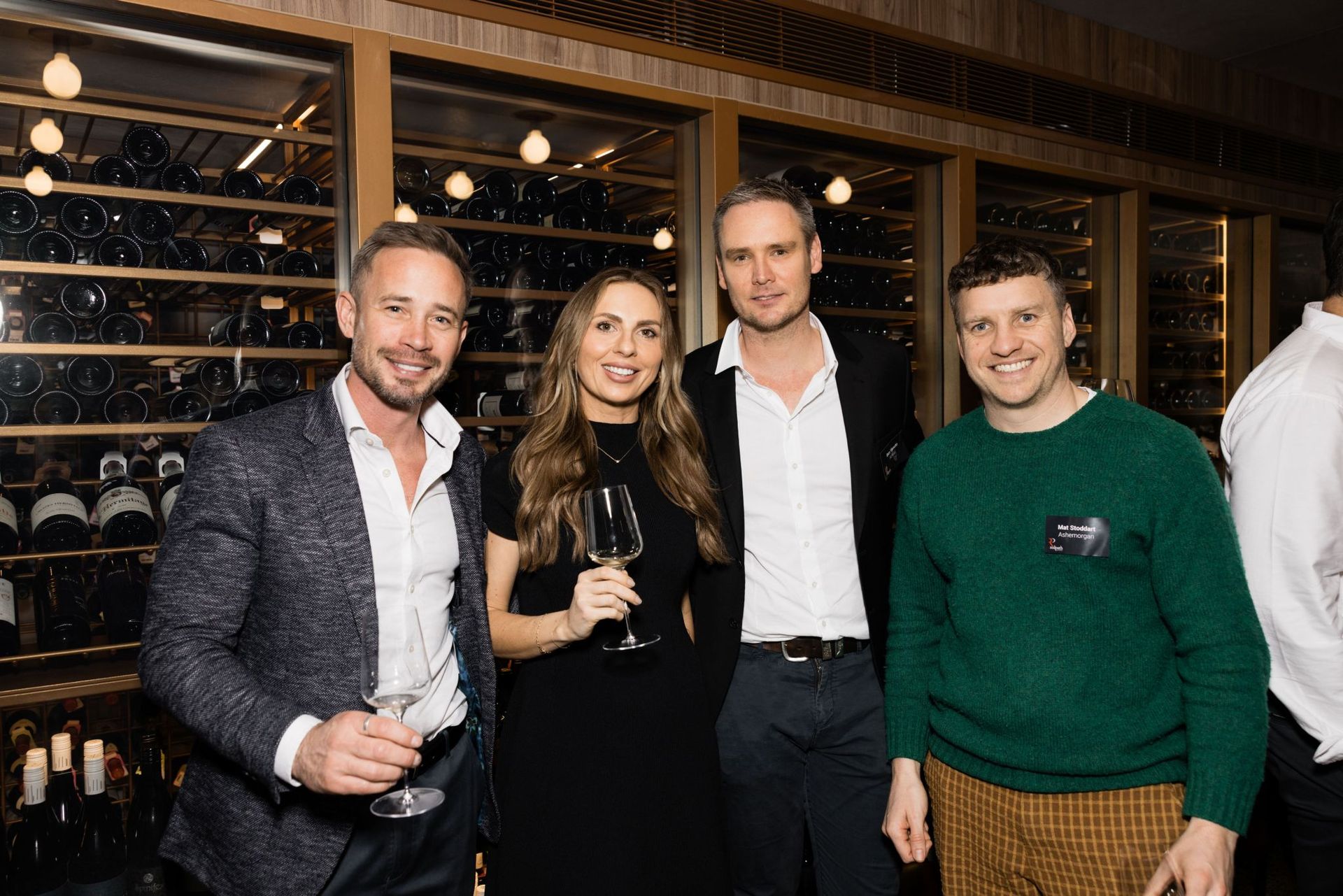 Four people pose for a photo in front of a wine rack, three holding glasses, smiling in a dimly lit, warm-toned setting.