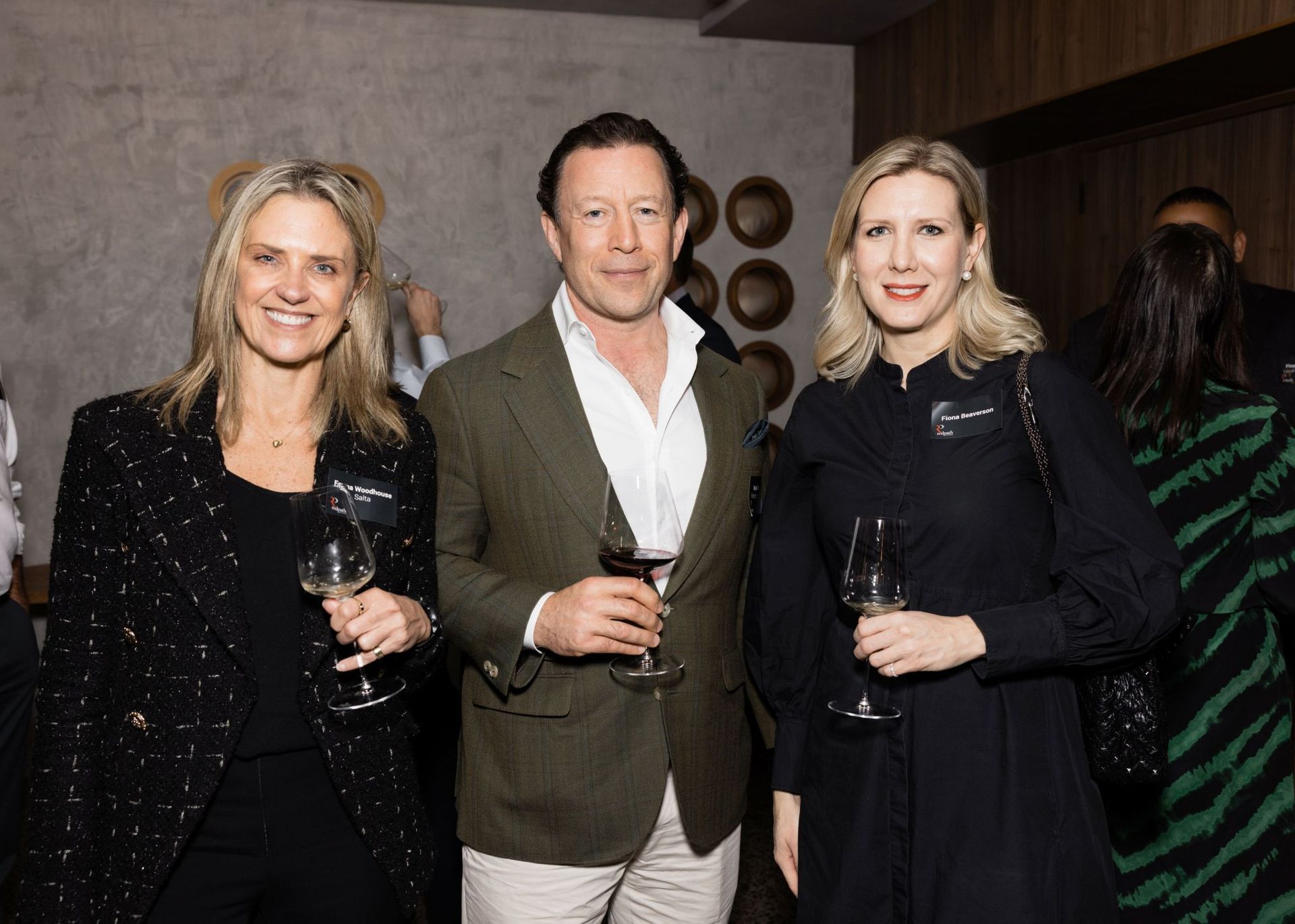 Three people smiling and holding wine glasses in an indoor social setting with a textured wall background.