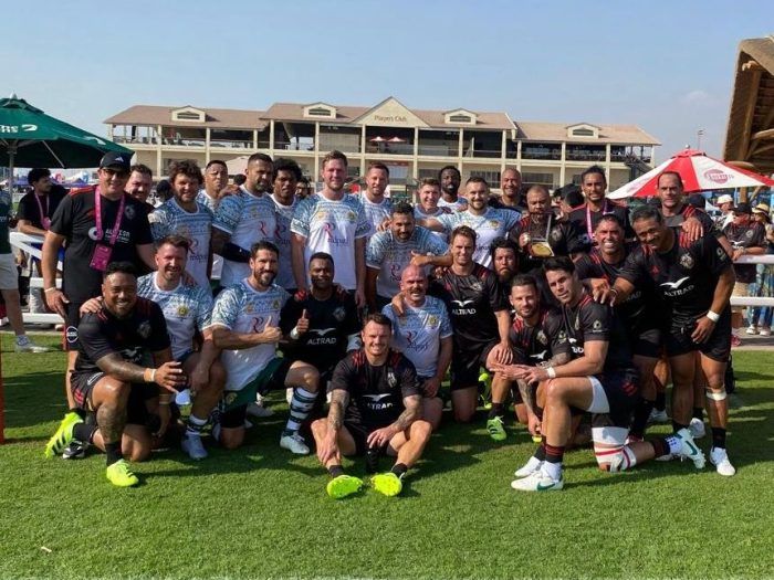A large group of rugby players in athletic uniforms posing for a team photo on a grassy field in front of a building.