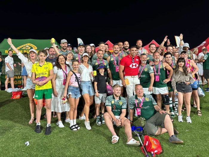 A diverse group of people celebrating on a field at night, some in rugby jerseys, standing and posing for a group photo.