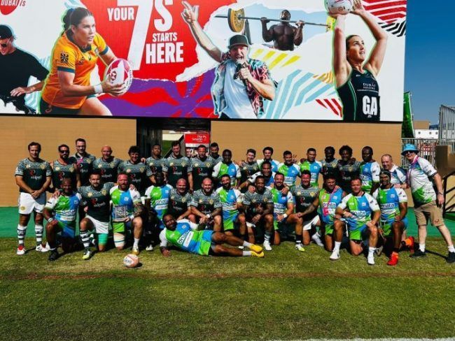 A large group of athletes in matching uniforms pose on a field in front of a colorful banner promoting local sports.