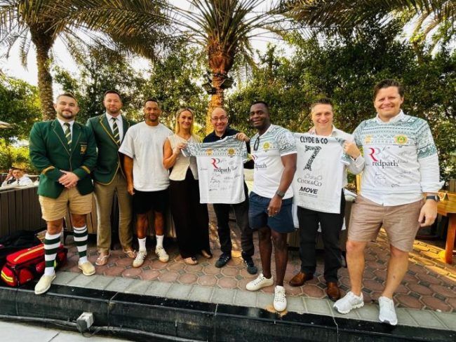 A group of eight people stand outdoors in a park setting, holding a white sports jersey for a photo.