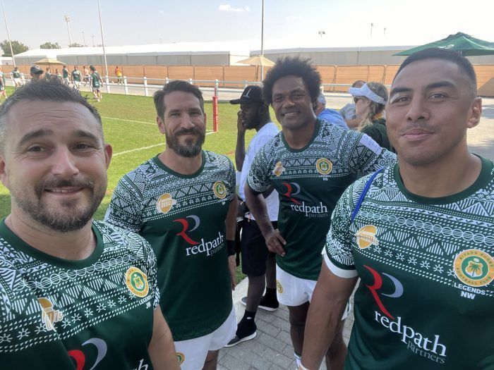 Four athletes in green and white patterned team uniforms with red logos pose for a selfie on an outdoor sports field.