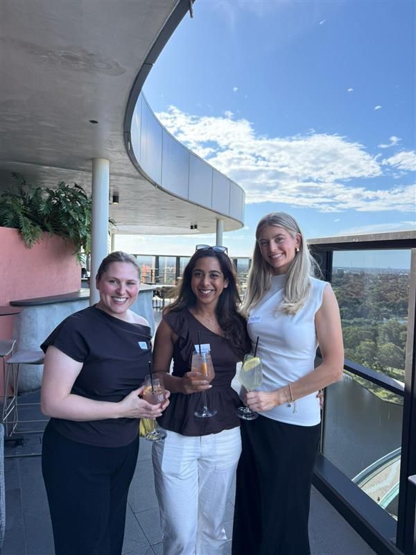 Three people smile while holding drinks on a rooftop terrace overlooking a river and bridge on a sunny day.