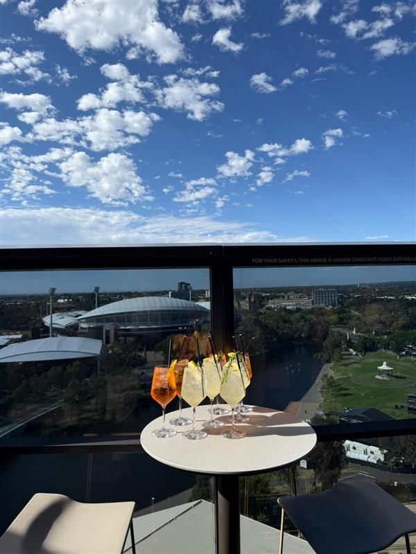 A round table with several cocktail glasses sits on a balcony overlooking a stadium, park, and blue sky with clouds.