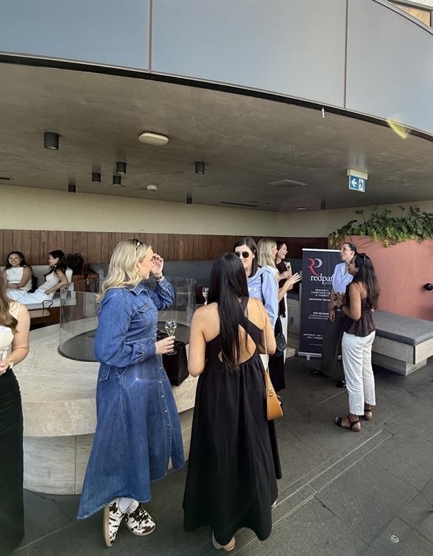 A group of people standing on an outdoor balcony at a social gathering near a branded event sign.