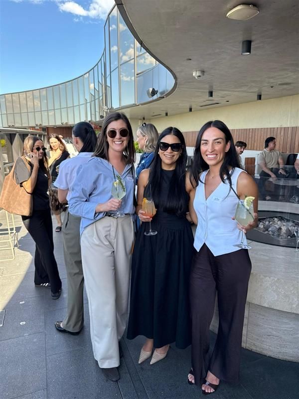 Four people posing with drinks on an outdoor terrace, wearing casual and formal attire with sunglasses.