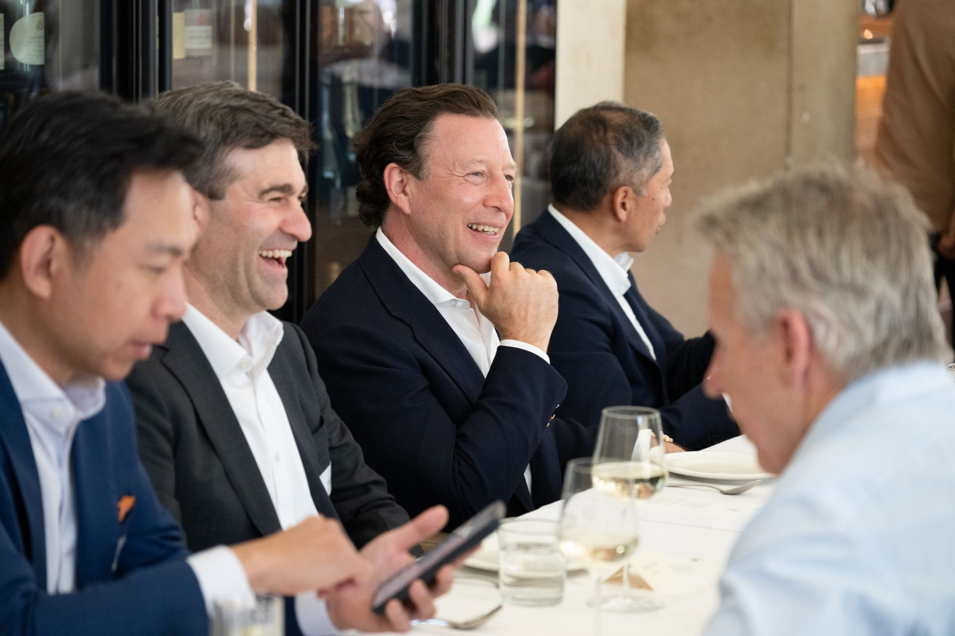 Five people in business attire laughing at a restaurant table, with one person in the foreground holding a smartphone.
