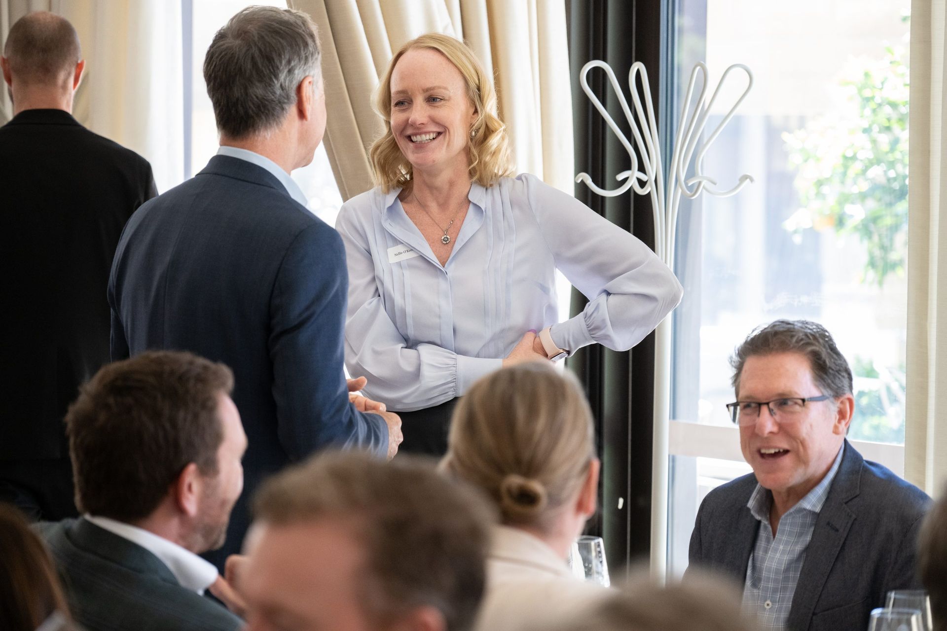 A group of professionals in business attire conversing and smiling in a well-lit, informal meeting space.