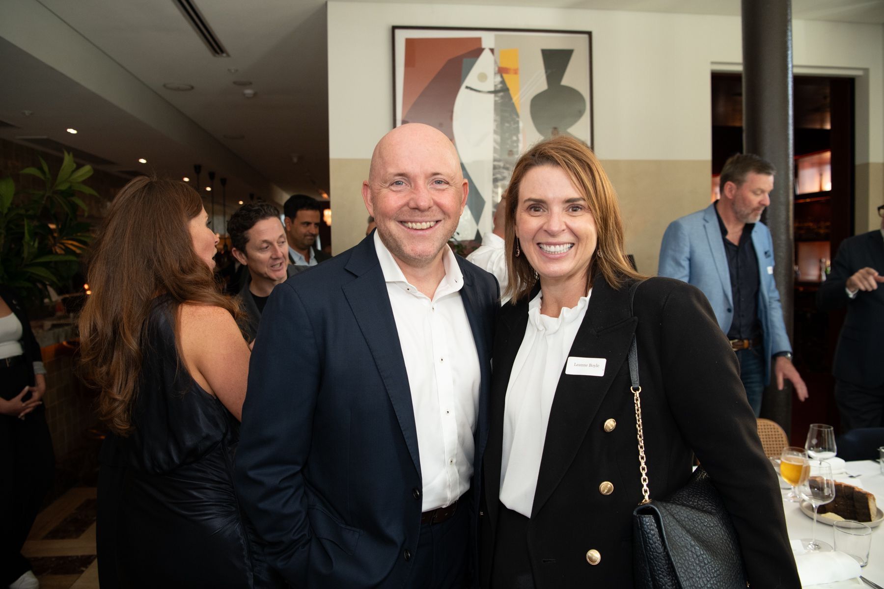 Two people smiling together in a room at a social gathering, with others blurred in the background.