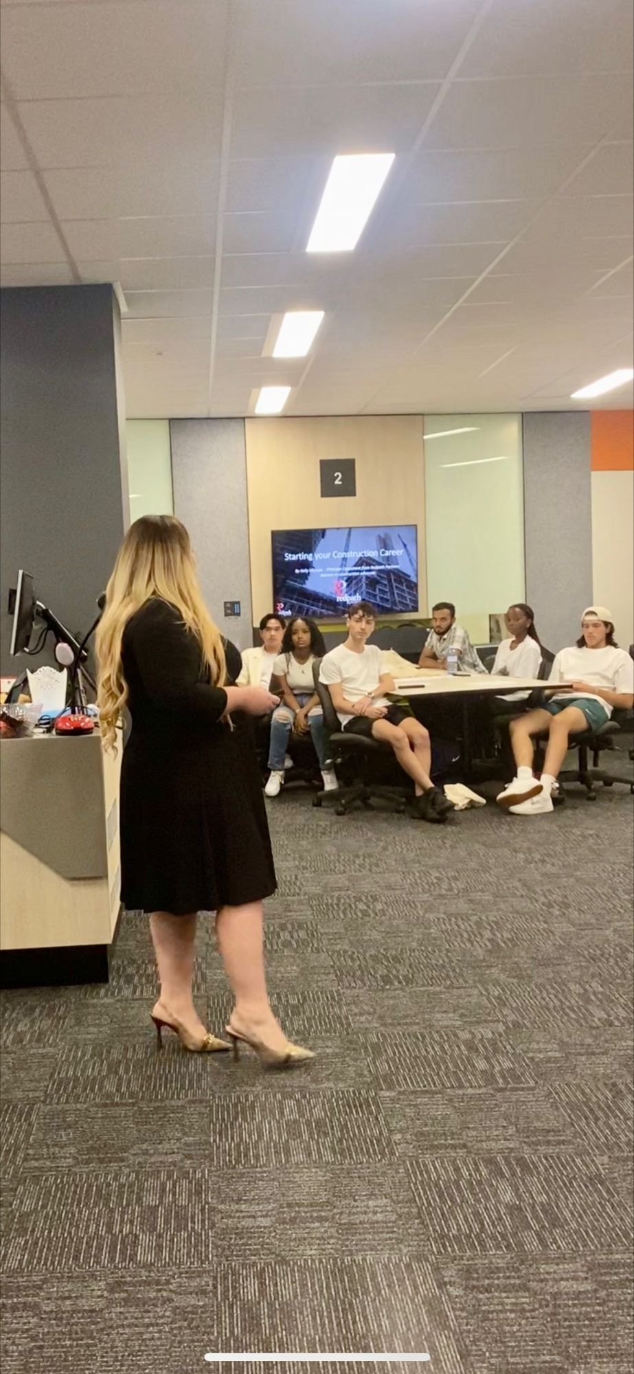 A speaker in a black dress stands before a group of people seated around a table in a brightly lit office or classroom.