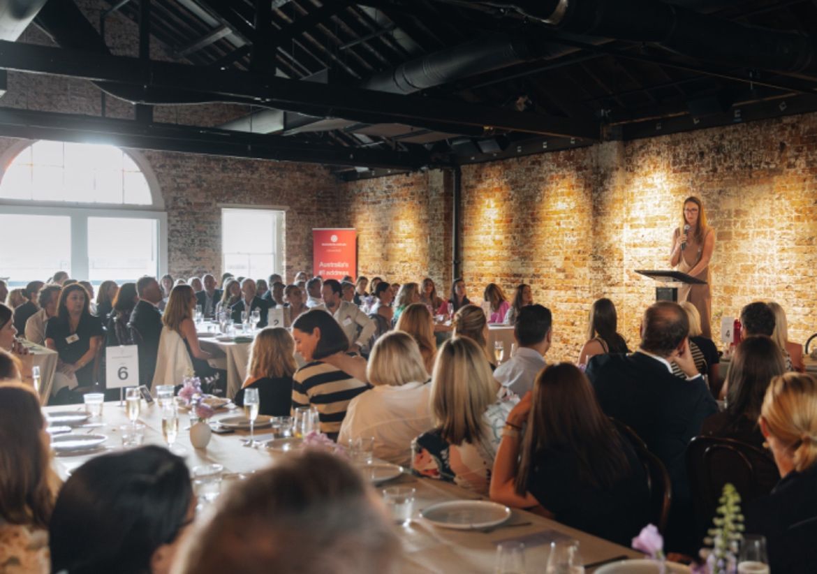 A speaker addresses a large audience seated at long tables in a rustic, brick-walled event space.