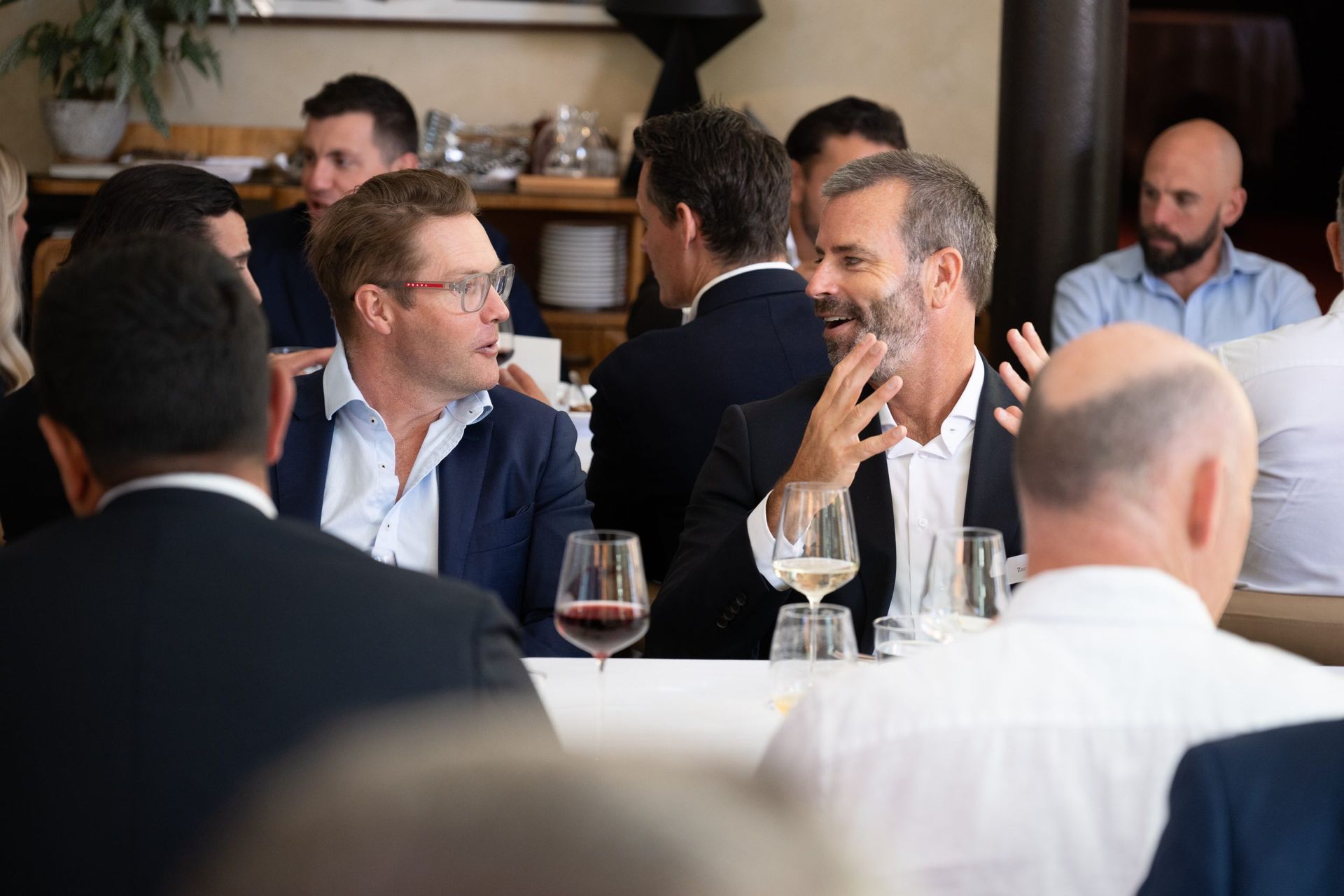 People in professional attire sit at a table in a restaurant, engaged in conversation and drinking wine.