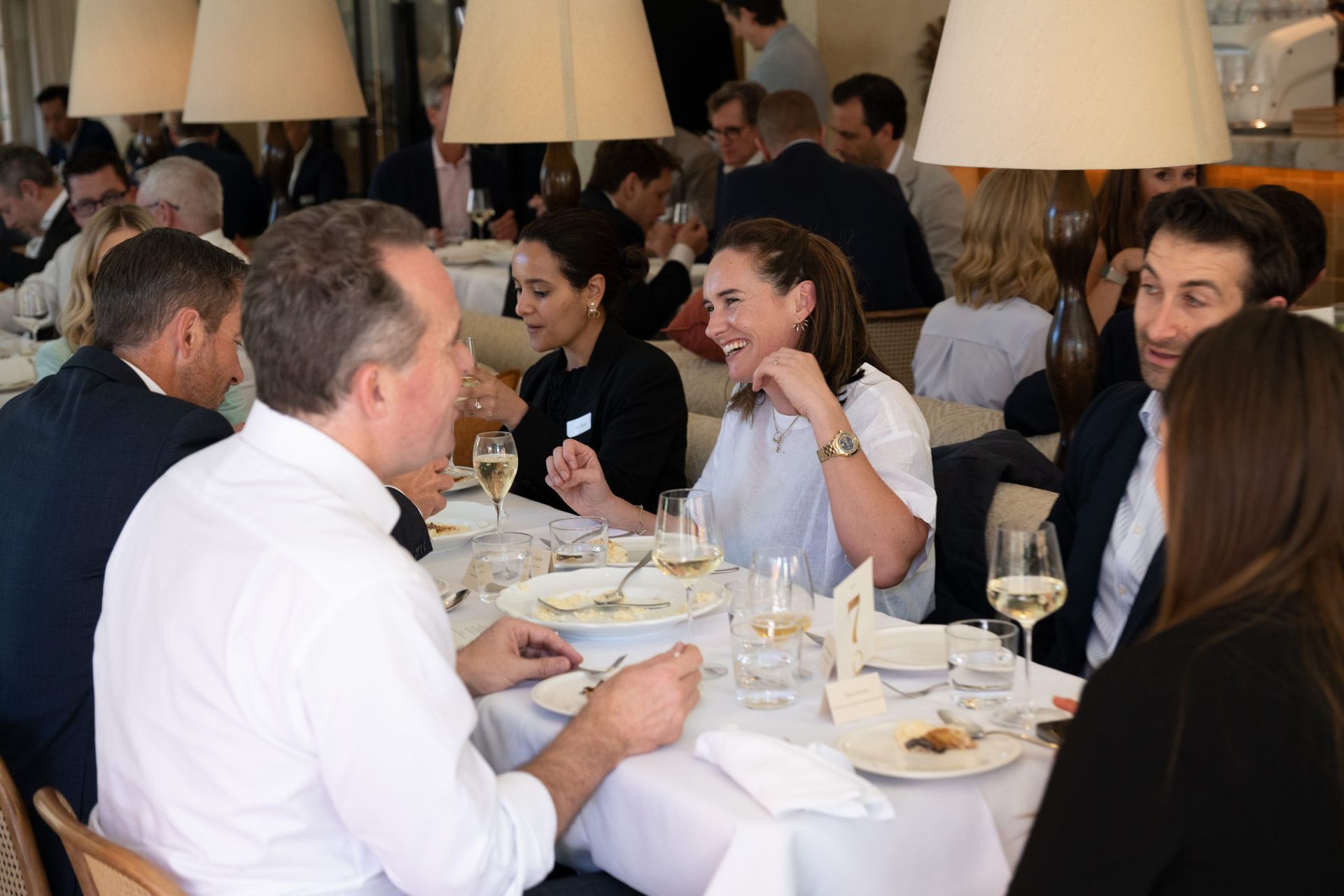 A lively lunch gathering where people sit around a long, white-clothed table in a well-lit dining area.