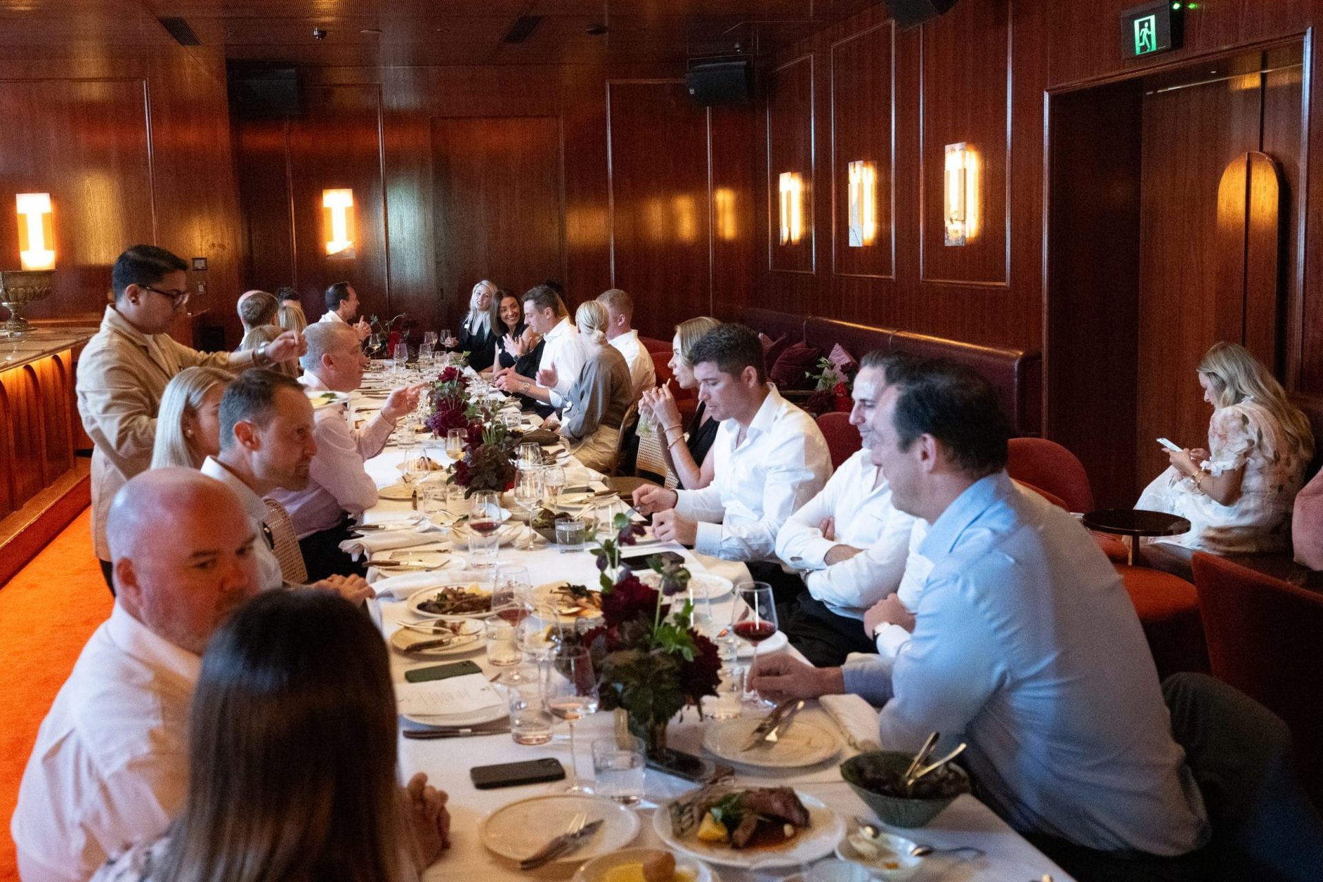 A group of people sits at a long table in a wood-paneled, dimly lit dining room during a formal dinner.