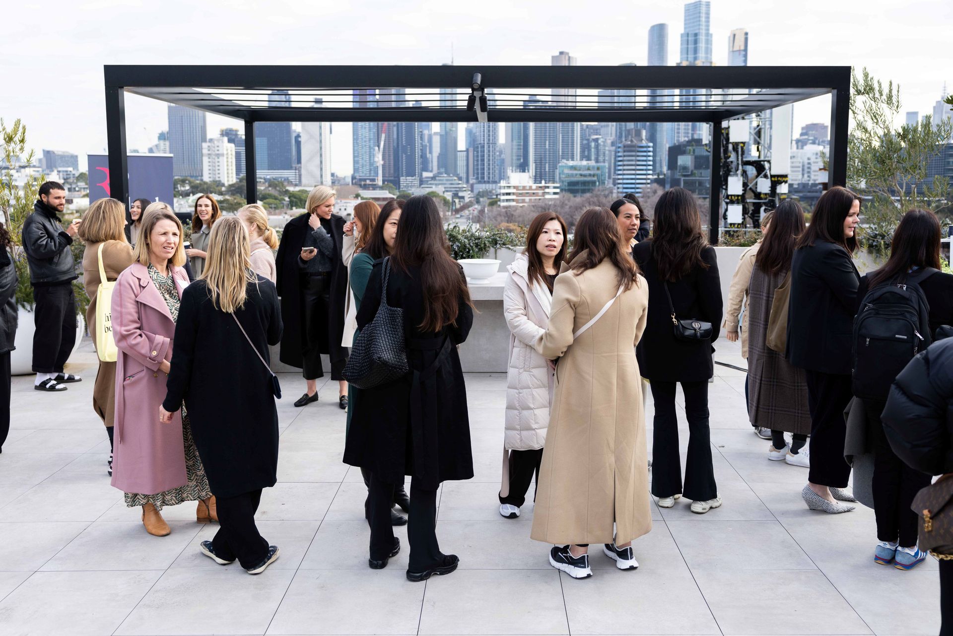 People stand and converse on a rooftop terrace with a city skyline visible in the background under a metal pergola.