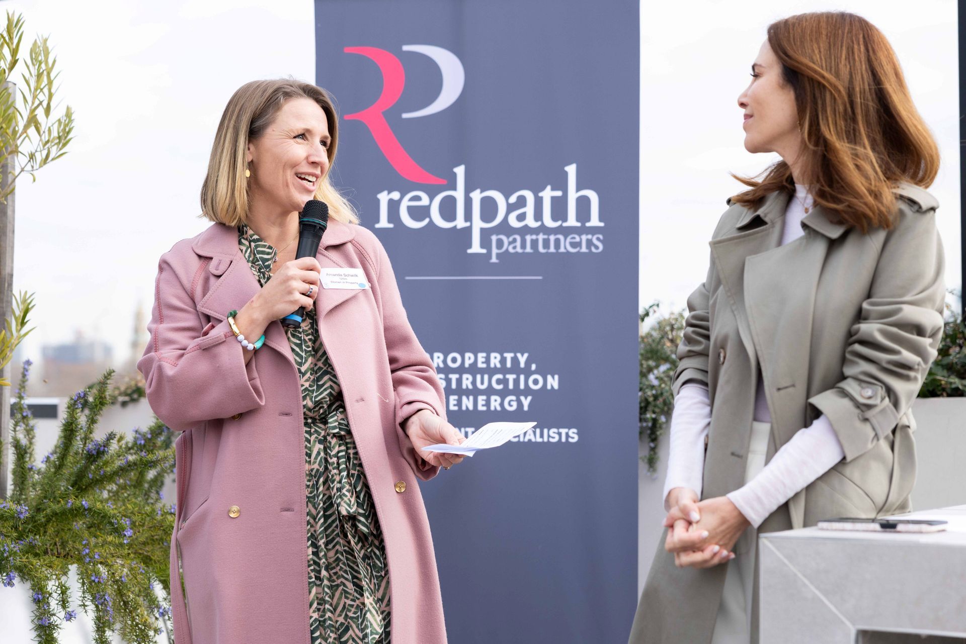 Two people stand at an outdoor event near a Redpath Partners banner, one speaking into a microphone, the other listening.
