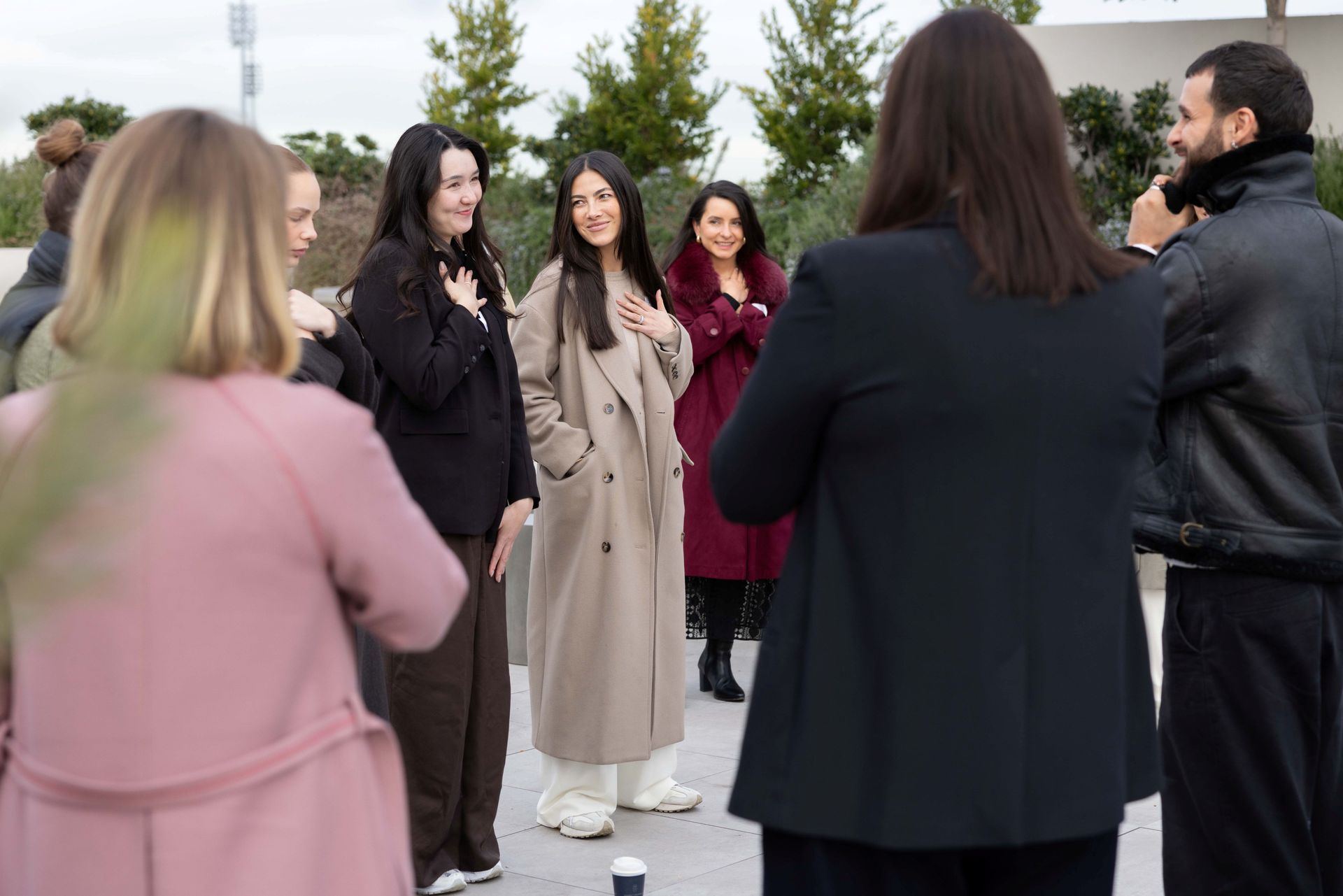 A group of people in stylish coats and blazers stand in a line outdoors, some holding their hands to their chests.