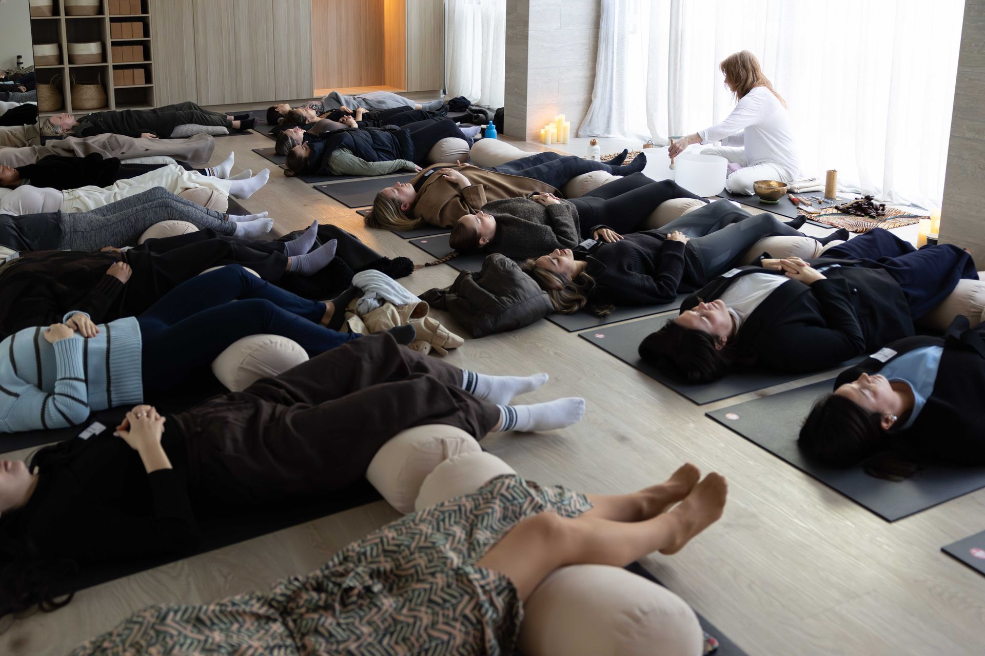 People lying on mats in a bright studio during a sound bath meditation session, with a facilitator nearby.