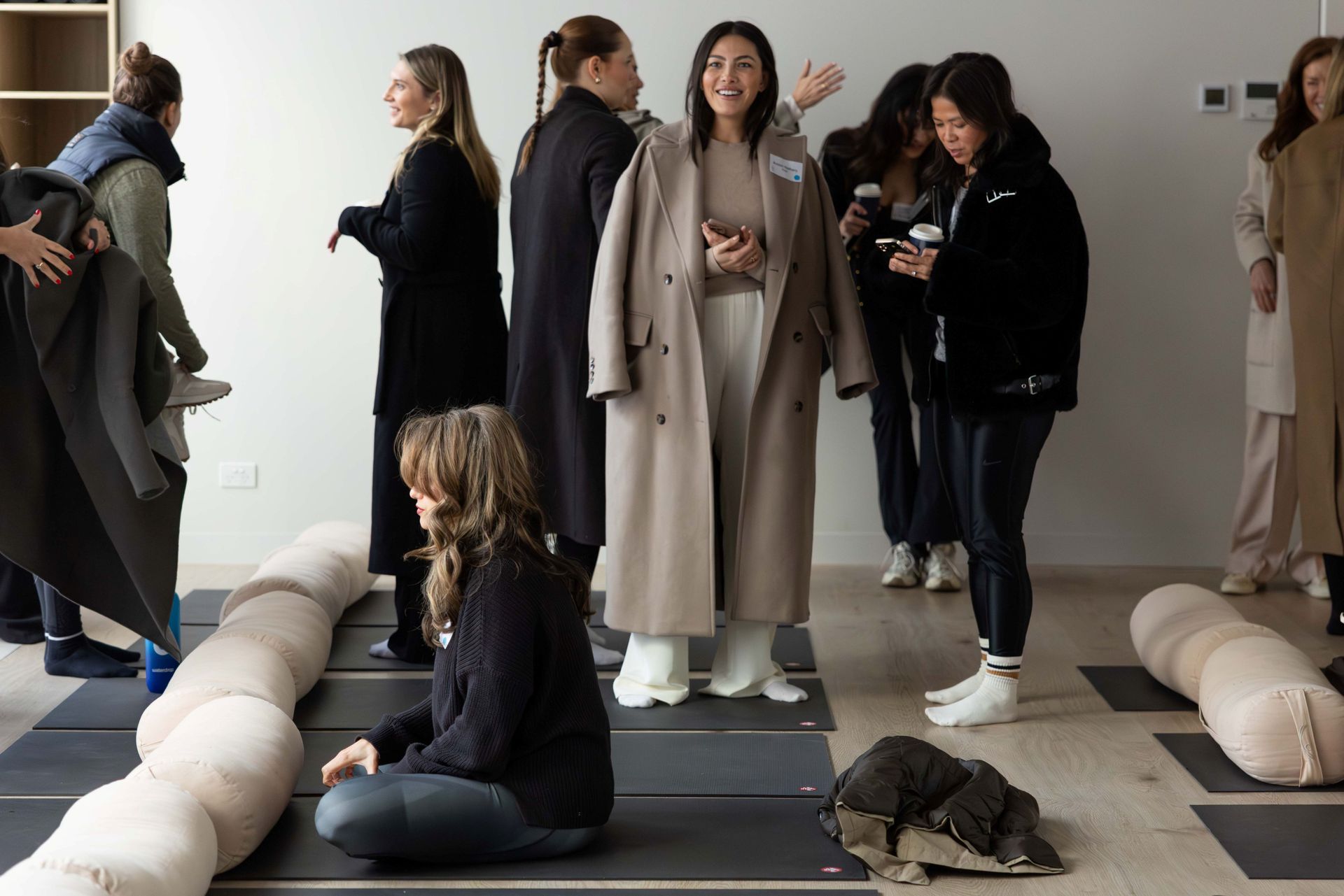 A group of people gathers in a bright studio, with some seated on yoga mats near beige bolsters and others standing and talking.