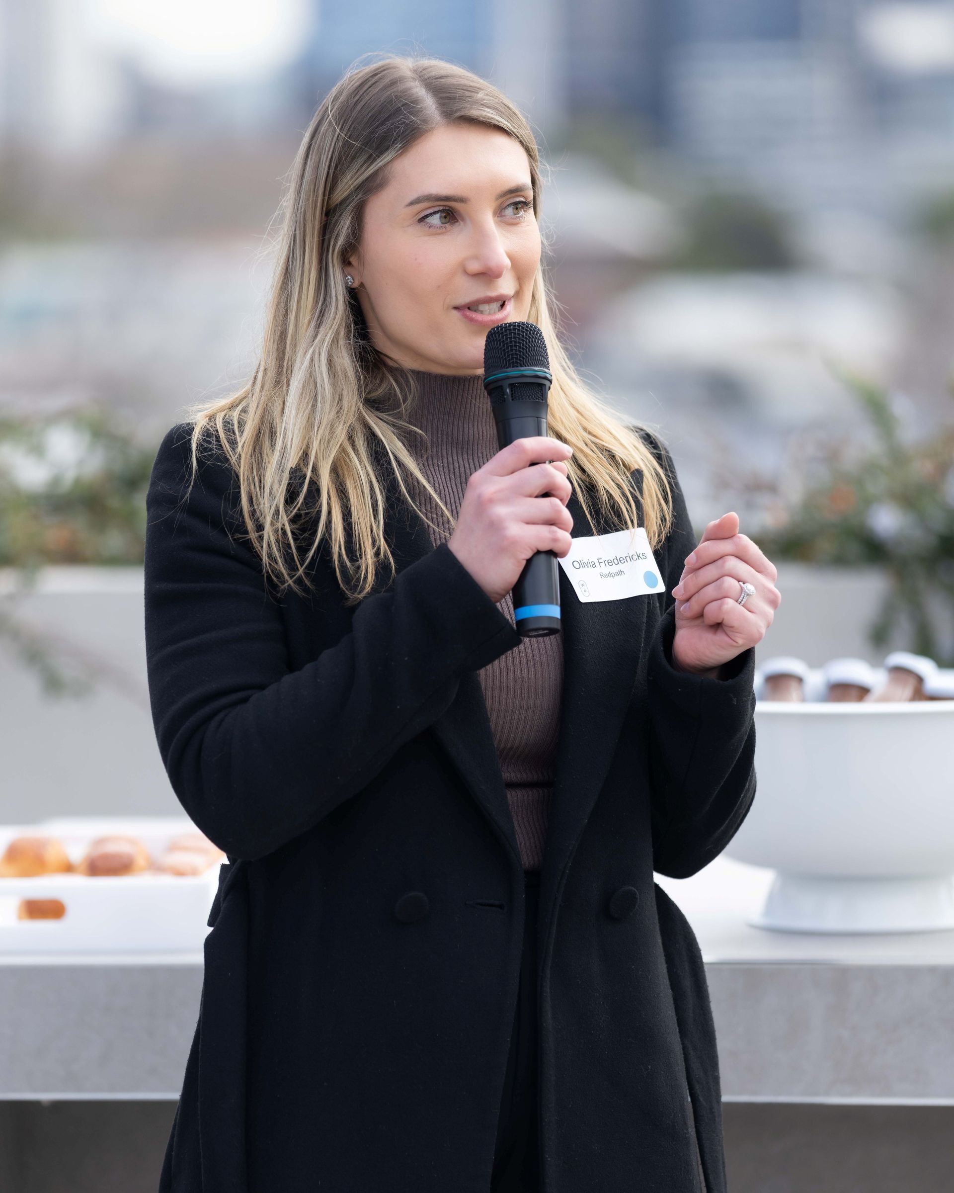 A person with long hair wearing a black coat speaks into a microphone at an outdoor event with a name tag attached.