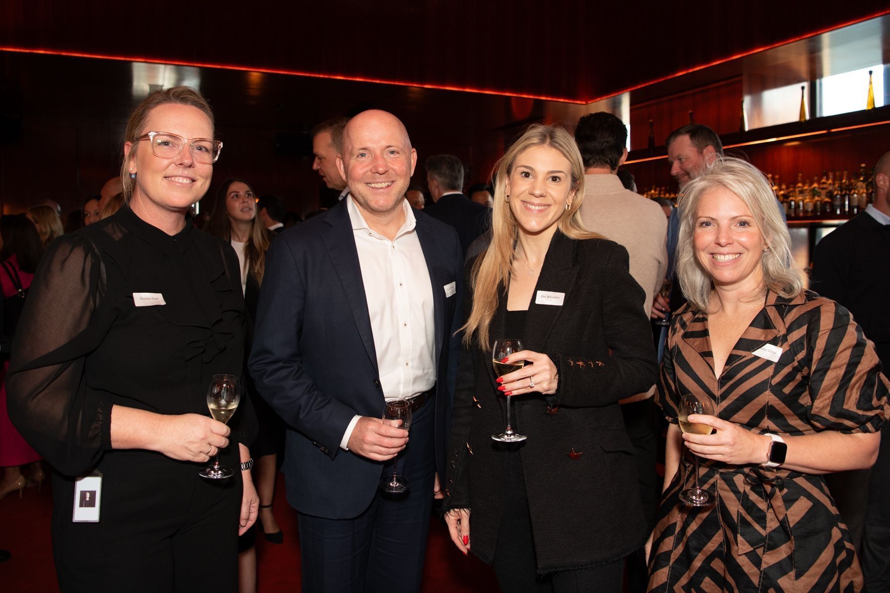 Four people holding drinks and smiling at a social gathering in a dimly lit, upscale venue with red lighting.