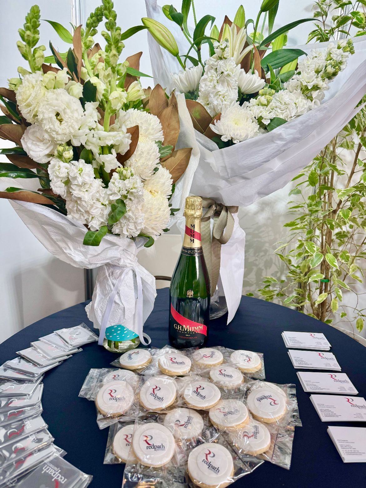 Two bouquets of white flowers and a bottle of champagne on a table with branded cookies and event cards.