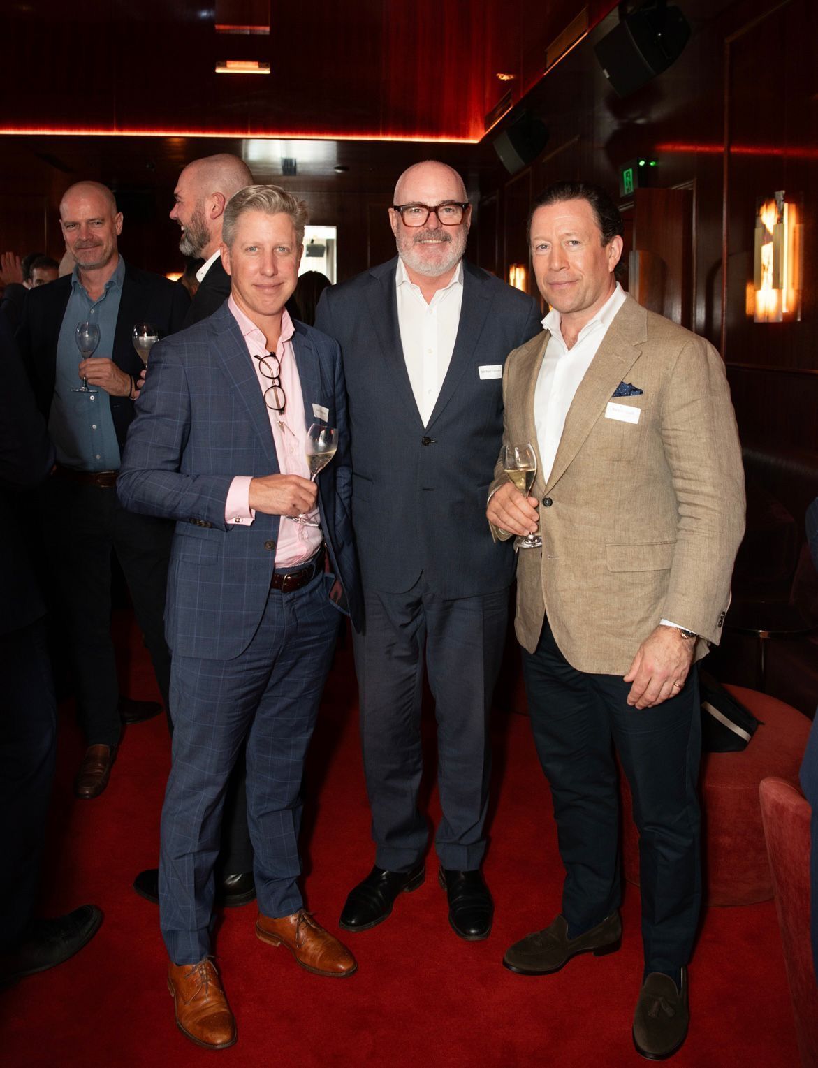 Three men in suits standing together in a dimly lit, red-carpeted lounge, posing for a photo while holding drinks.