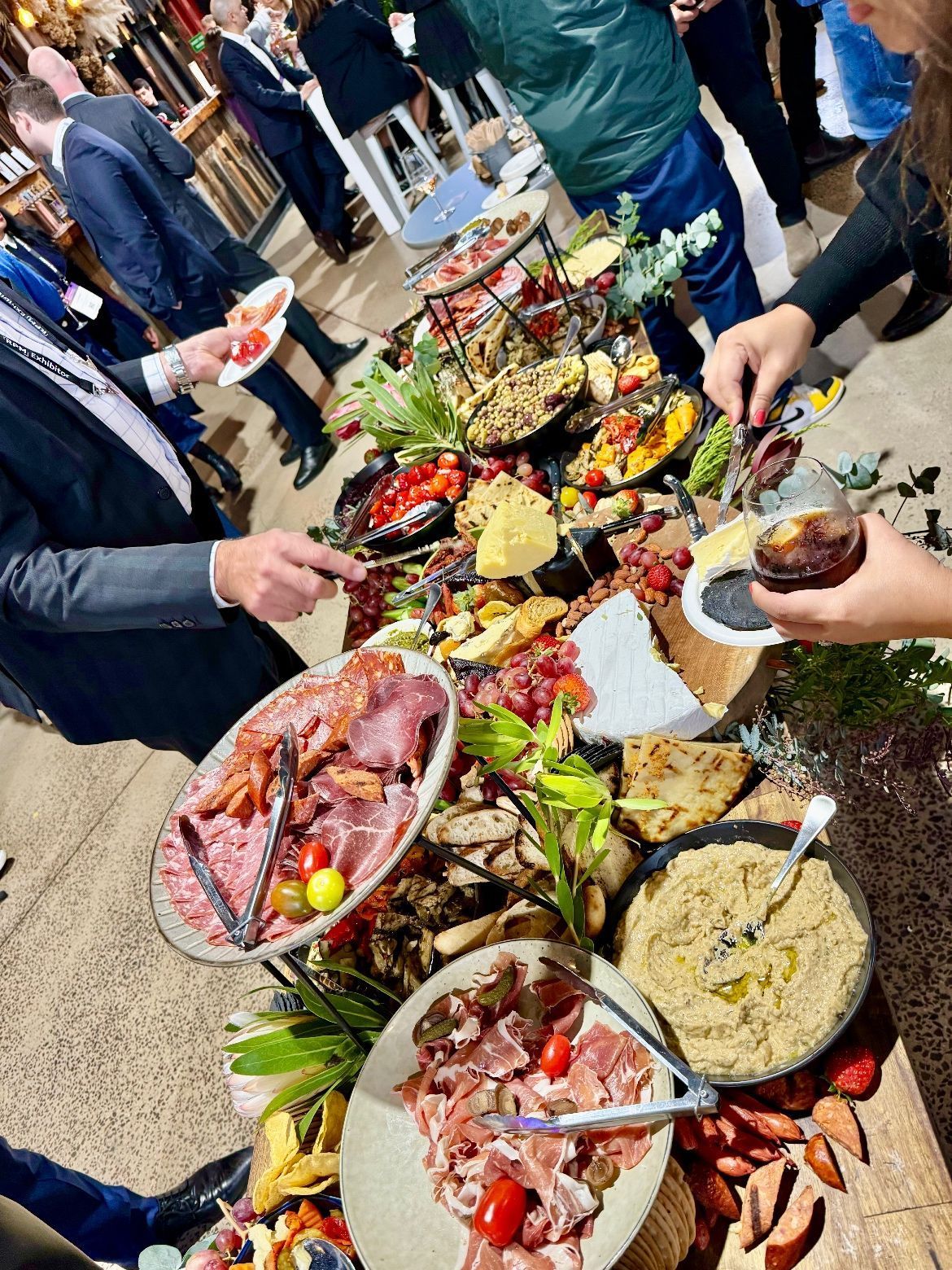 A high-angle view of a crowded buffet table filled with cured meats, appetizers, and dips, with guests serving themselves.