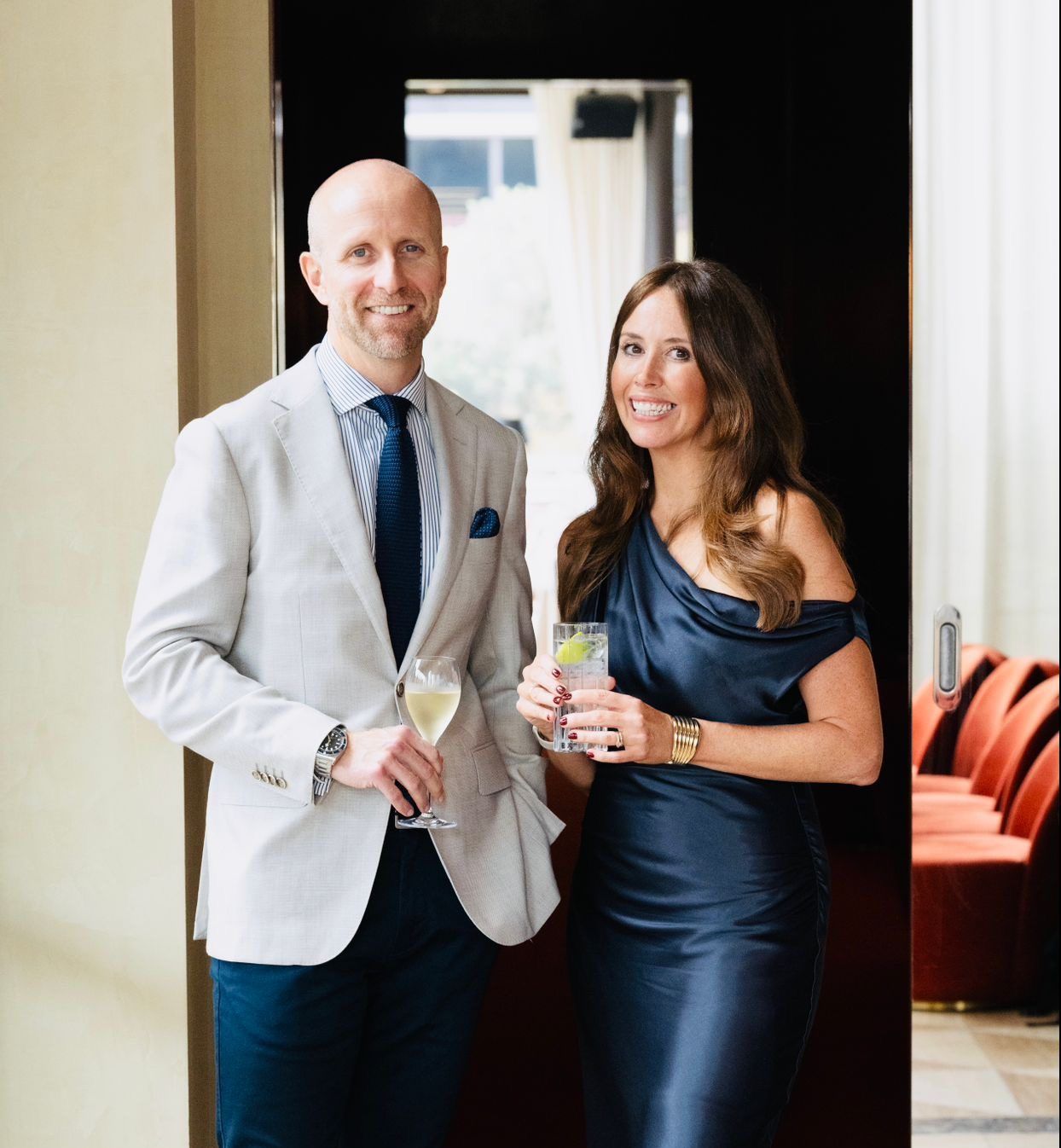 A man in a light blazer and a woman in a navy dress stand together holding drinks in a modern, well-lit interior.