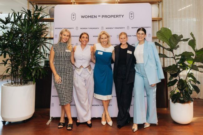 Five people stand side-by-side in front of a Women in Property backdrop in a brightly lit, professional indoor setting.
