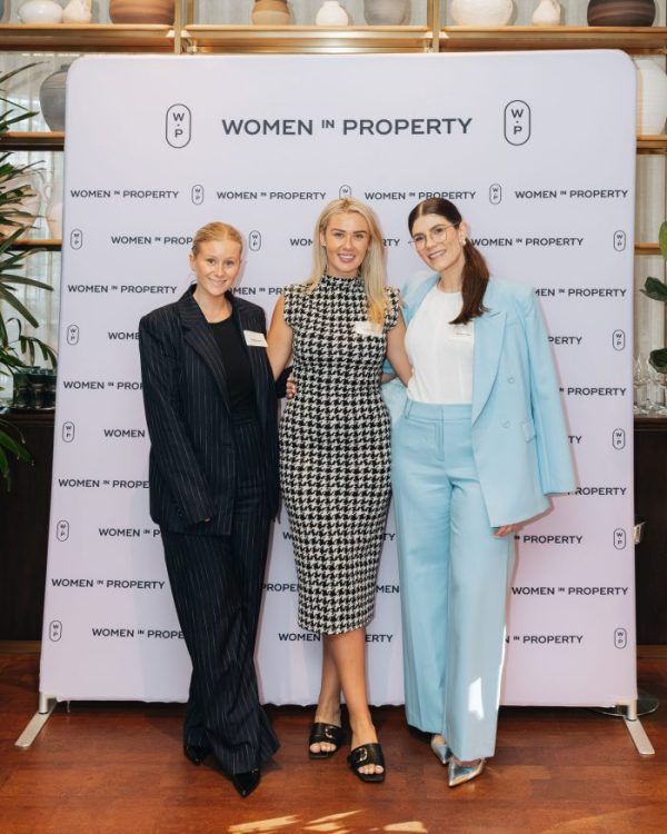 Three people stand together smiling in front of a branded Women in Property backdrop at an indoor event.