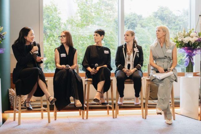 Five people seated in a row on high chairs at an indoor panel discussion, holding microphones and facing an audience.